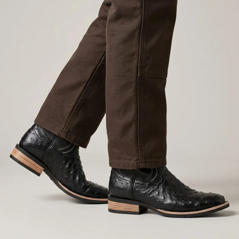 Yehaw CHARRO black vegan leather cowboy boots with textured ostrich-style finish and low heel worn with brown trousers.