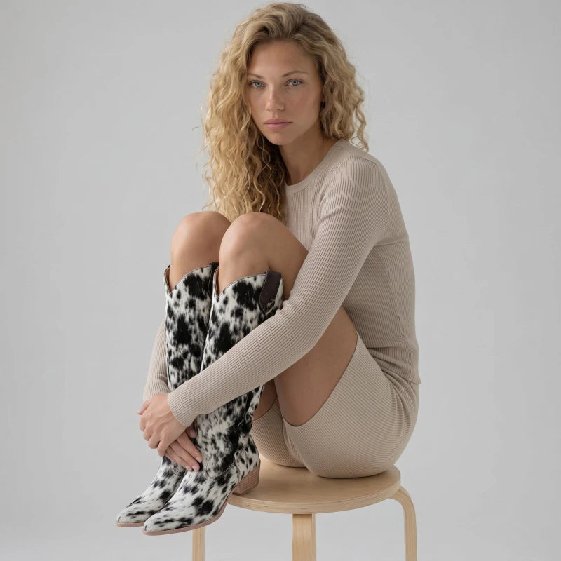 Model wearing Yehaw MILK knee-high black and white cow print vegan suede cowboy boots with a pointed toe.