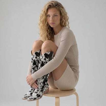 Model wearing Yehaw MILK knee-high black and white cow print vegan suede cowboy boots with a pointed toe.